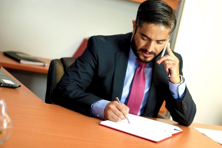 Businessman in a dark suit writes in a notebook at a desk while talking on a mobile phone held to his ear.