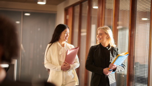 Two professional women walk and chat in a sunlit office hallway, each carrying folders and smiling at each other, one in a beige suit and the other in a black blazer.