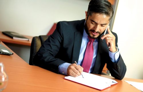 Businessman in a dark suit writes in a notebook at a desk while talking on a mobile phone held to his ear.