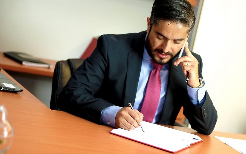 Businessman in a dark suit writes in a notebook at a desk while talking on a mobile phone held to his ear.