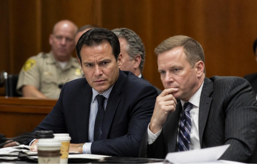Two men in dark suits sit at a wooden table, serious faces, coffee cups and papers in front.