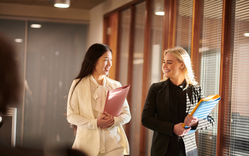 Two professional women walk and chat in a sunlit office hallway, each carrying folders and smiling at each other, one in a beige suit and the other in a black blazer.