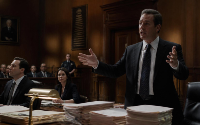 A man in a suit stands and gestures while speaking in a wood-paneled courtroom, with colleagues seated at a desk piled high with legal documents in front of him.
